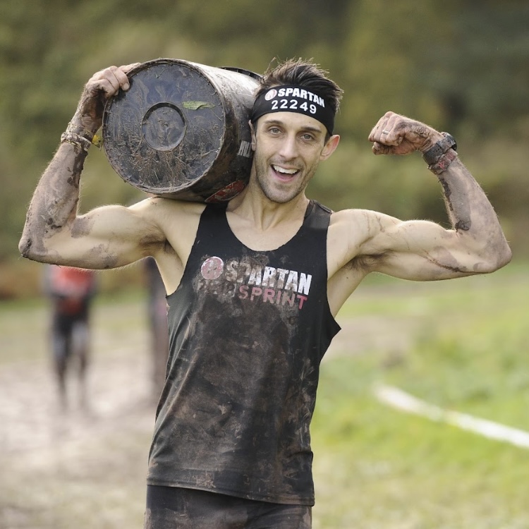 White male with brown hair, smiling flexing biceps while carrying a barrel and covered in mud