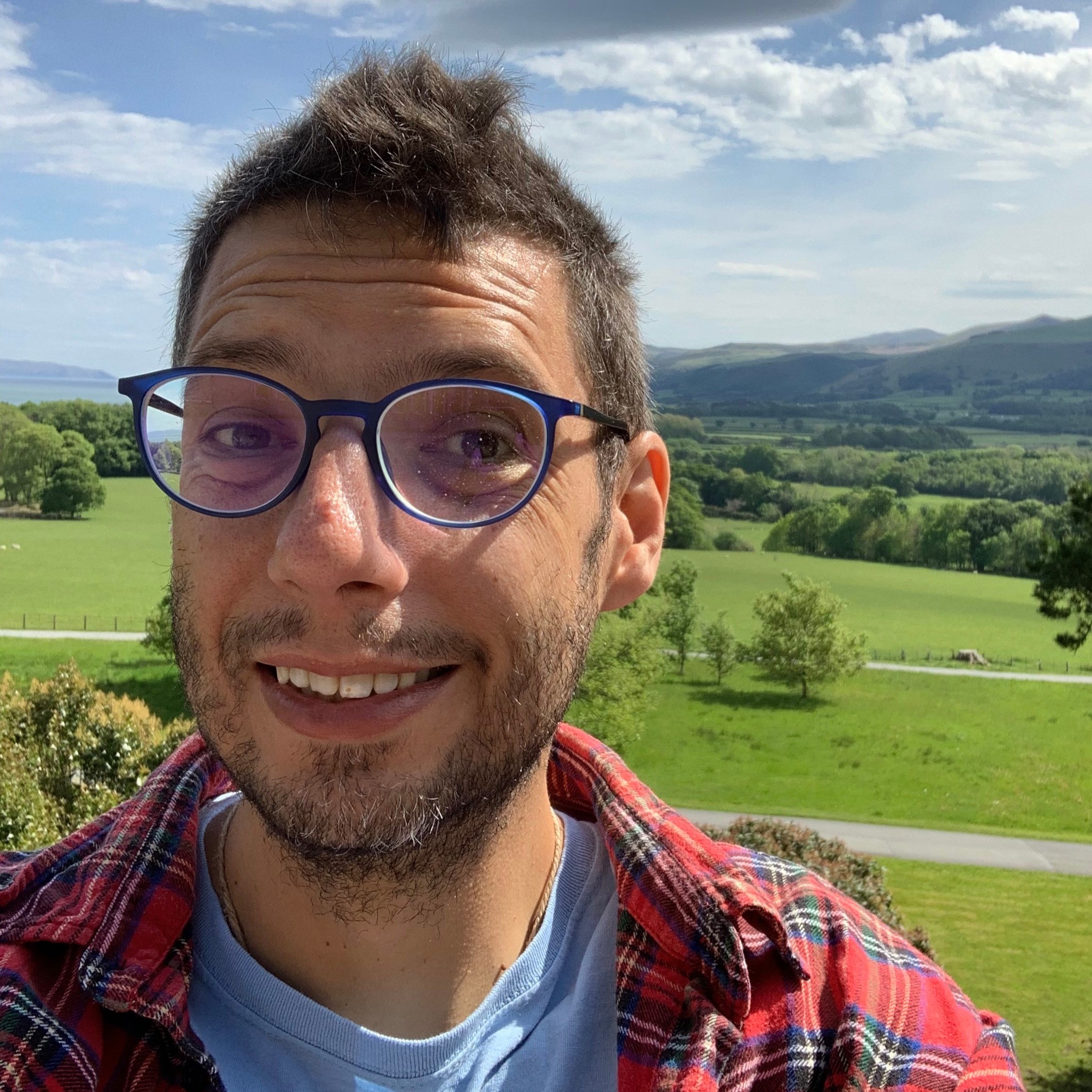 Mark with glasses smiling, brown hair, blue sky and green hills in background, red shirt