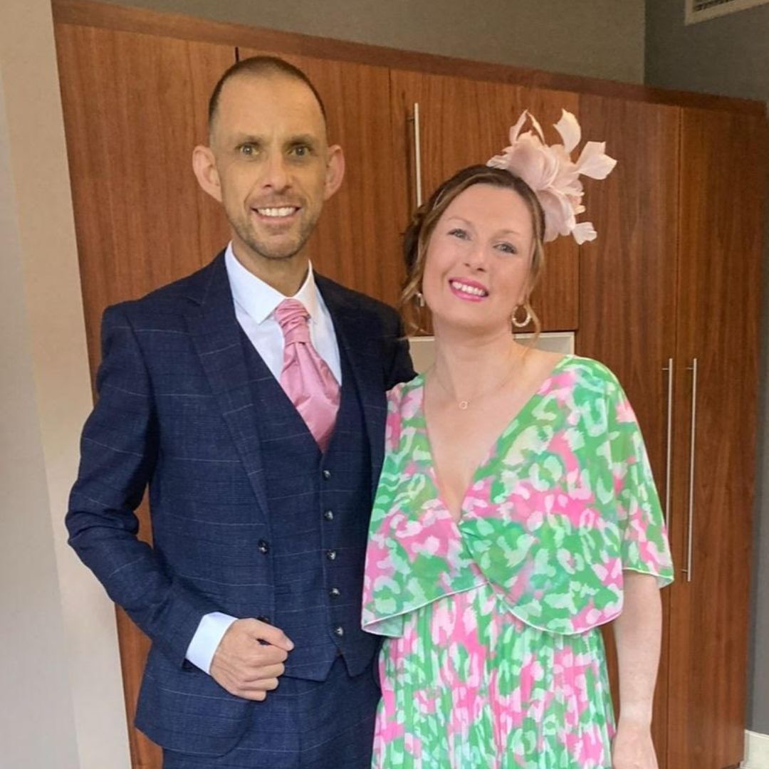 imgKT20250507 (1) Man on left in dark blue suit, pink tie and white shirt with jaundiced skin, smiling at camera. Woman on right in green and pink floral dress and pink fascinator, smiling at camera.