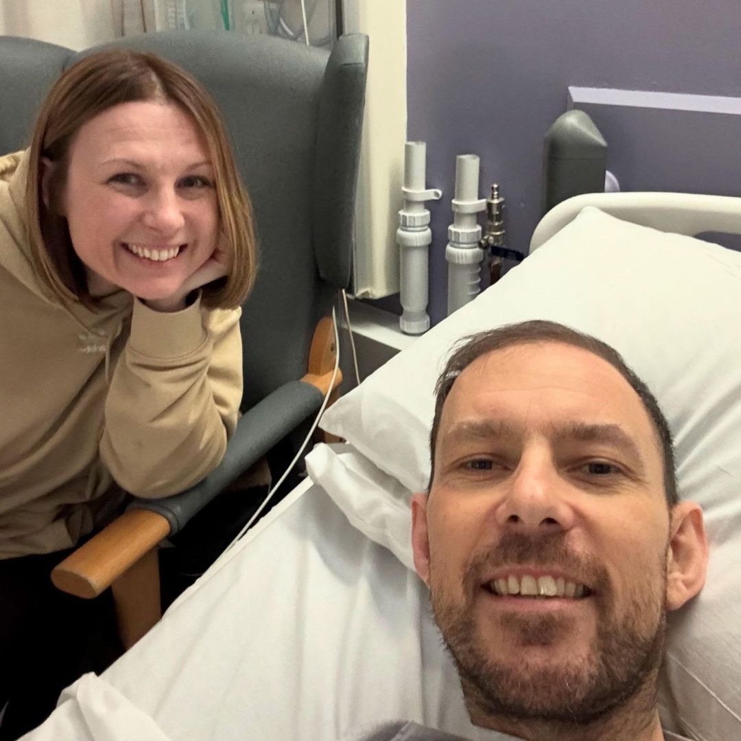 imgKT20250507 Woman top left in light brown hoodie smiling with hand on chin, man in foreground in hospital bed smiling at camera with slightly jaundiced skin