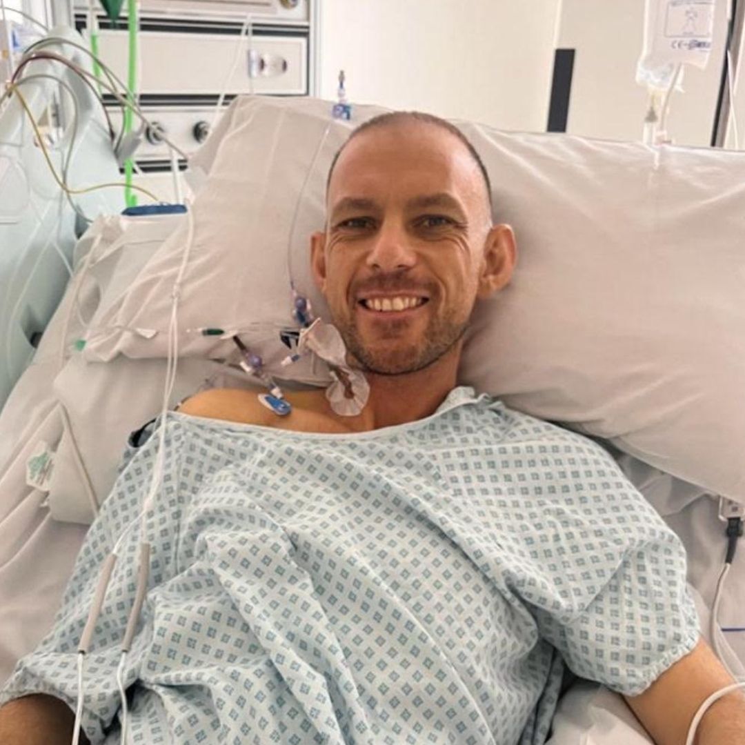 imgKT20250507v3 (1) Man in hospital bed angled sitting up, smiling at camera with tubes coming from his neck.