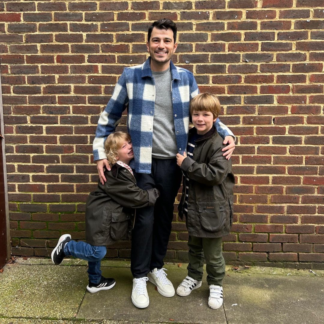 Father with light skin and brown hair with arms around his two young sons stood just in front of him with a brick wall background