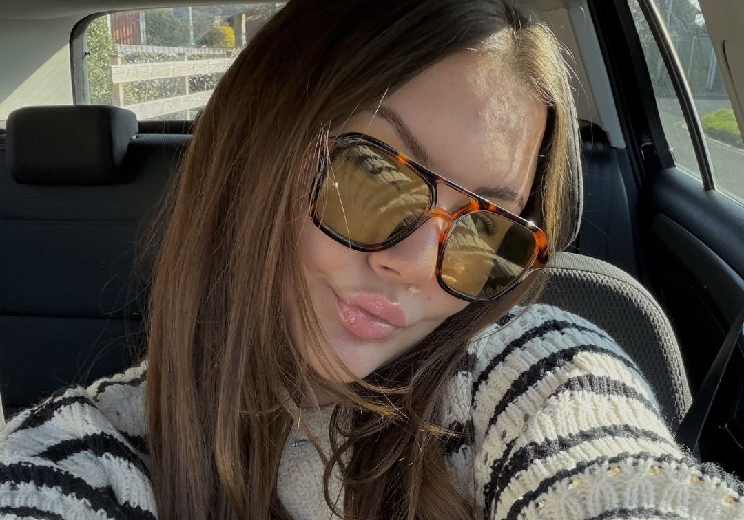 Amy in her car with sunglasses on pouting to camera; long brown hair, sun on her face